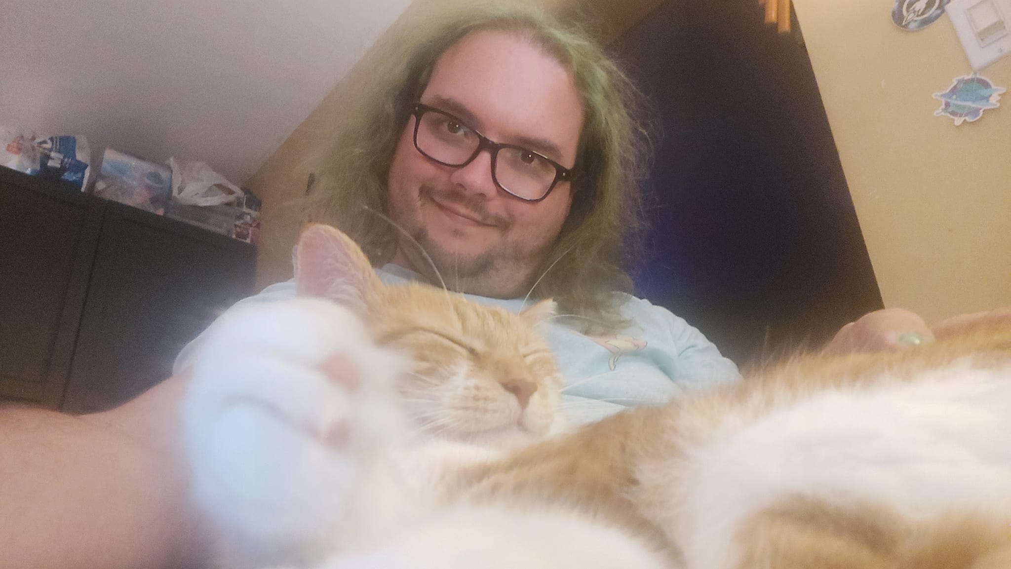 A man with long green hair and no beard holds an orange cat who is sleeping contentedly